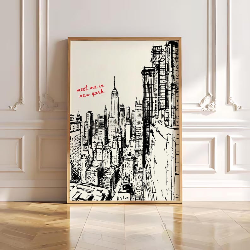 May include: A framed black and white sketch of the New York City skyline, including the Empire State Building. The words "meet me in new york" are written in red script. The artwork is displayed in a room with white walls and a wooden floor.