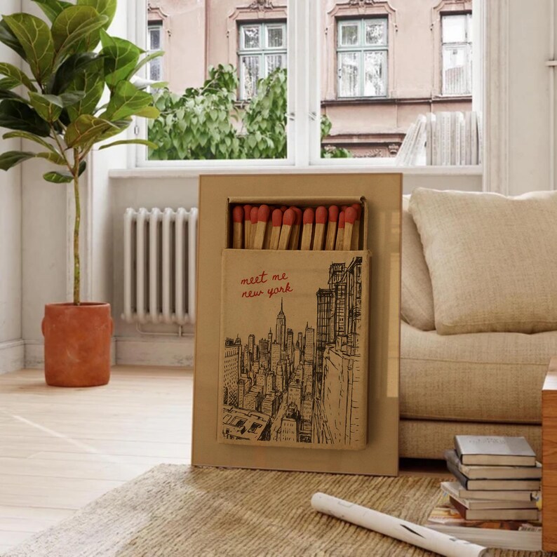 Puede incluir: Una caja de cerillas rectangular con una ilustraci&oacute;n del horizonte de la ciudad de Nueva York y el texto "meet me new york". La caja de cerillas est&aacute; llena de cerillas con punta roja. La caja de cerillas est&aacute; colocada en un marco de cart&oacute;n m&aacute;s grande.