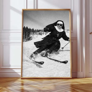 Könnte beinhalten: Gerahmter Schwarz-Weiß-Druck einer Nonne, die eine schneebedeckte Piste hinunterfährt. Die Nonne trägt eine Tracht, eine Sonnenbrille und ein Kreuz. Das Bild ist eine Vintage-Fotografie.
