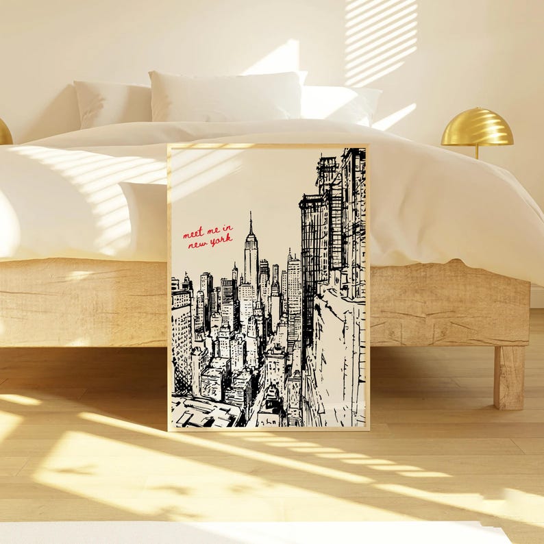 May include: A framed black and white illustration of the New York City skyline, featuring the Empire State Building. The words "meet me in new york" are written in red script. The artwork is displayed in a bedroom setting.