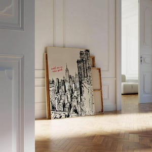 May include: A framed canvas print featuring a black and white illustration of the New York City skyline. The print includes the text "meet me in new york" in red script. The artwork is leaning against a white wall.
