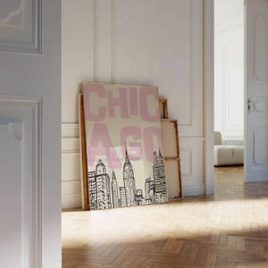 May include: A framed art print featuring a stylized black and white skyline of Chicago, with the word "CHICAGO" in pink block letters. The artwork is displayed against a neutral background, leaning against a wall.