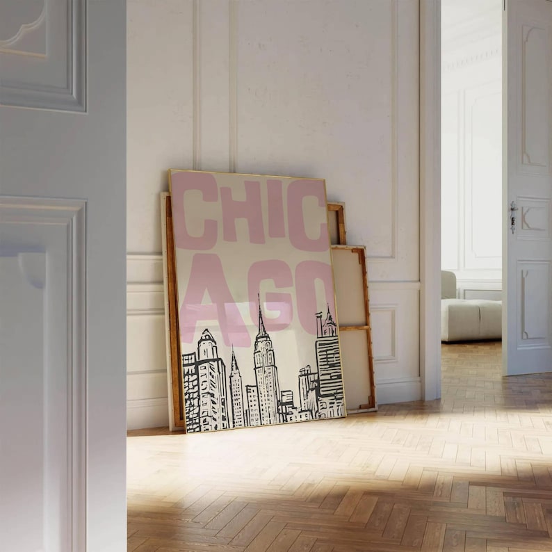 May include: A framed art print featuring a stylized black and white skyline of Chicago, with the word "CHICAGO" in pink block letters. The artwork is displayed against a neutral background, leaning against a wall.