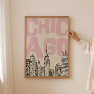 May include: A framed art print featuring a stylized skyline of Chicago in black and white. The word "CHICAGO" is printed in large, pink letters across the top. The artwork is displayed in a light-colored wooden frame.