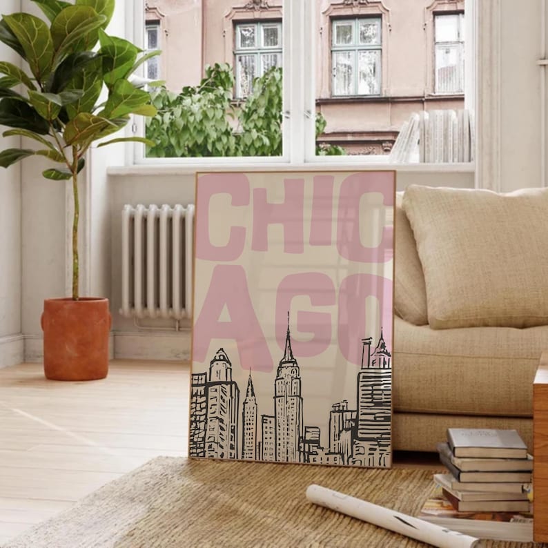 May include: A framed art print featuring the word "CHICAGO" in large pink letters over a beige background. Below the text is a black and white skyline illustration of Chicago's iconic buildings. The artwork is displayed in a room with natural light.