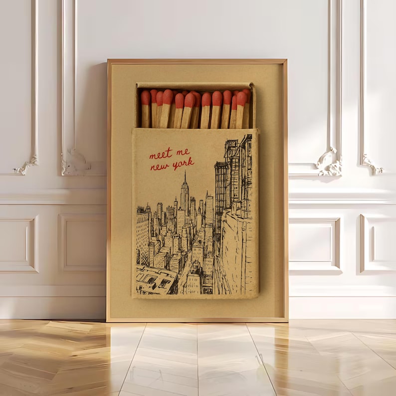 Puede incluir: Obra de arte enmarcada con un dise&ntilde;o de caja de cerillas que representa el horizonte de la ciudad de Nueva York. La caja de cerillas est&aacute; abierta, revelando cerillas con puntas rojas. El texto "meet me new york" est&aacute; escrito en rojo.
