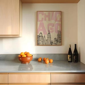 May include: A kitchen scene featuring a framed poster with the word "CHICAGO" in pink, above a countertop with a bowl of oranges and two wine bottles. The poster depicts a skyline. The bowl is terracotta.