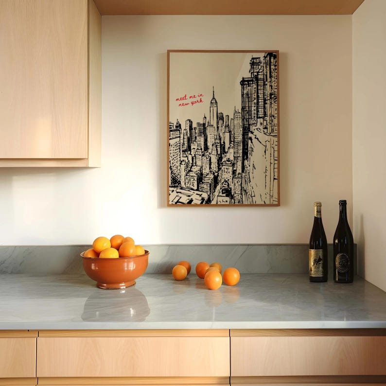 May include: A framed black and white illustration of the New York City skyline hangs on a white wall. The text "meet me in new york" is written in red. A terracotta bowl filled with oranges sits on a gray countertop, with two wine bottles to the right.
