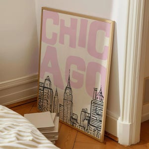 May include: A framed art print featuring the word "CHICAGO" in pink block letters over a black and white skyline illustration. The print is in a gold frame and leans against a white wall.