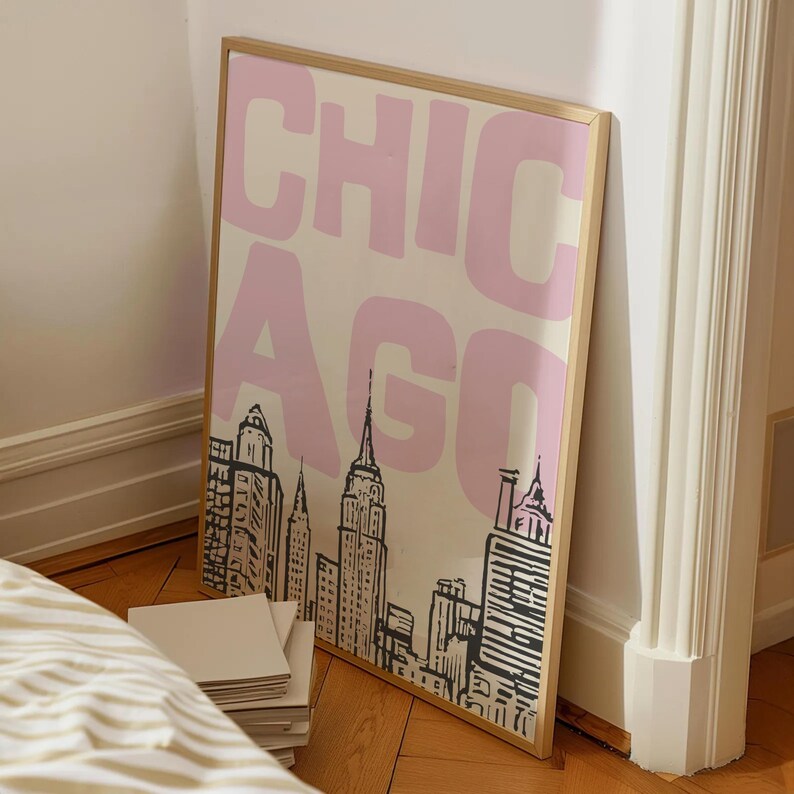 May include: A framed art print featuring the word "CHICAGO" in pink block letters over a black and white skyline illustration. The print is in a gold frame and leans against a white wall.