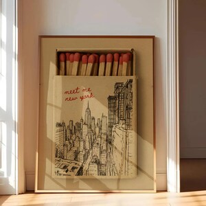 Puede incluir: Obra de arte enmarcada que presenta un dise&ntilde;o de caja de cerillas con una ilustraci&oacute;n en blanco y negro del horizonte de la ciudad de Nueva York. La caja de cerillas est&aacute; abierta, revelando cerillas con puntas rojas. El texto "meet me new york" est&aacute; escrito en rojo.