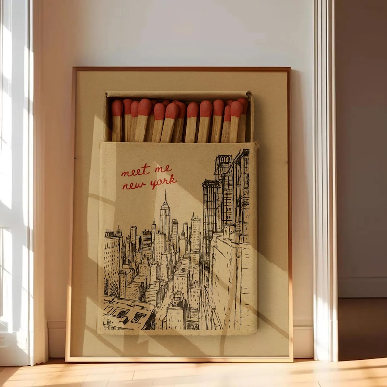 Puede incluir: Obra de arte enmarcada que presenta un dise&ntilde;o de caja de cerillas con una ilustraci&oacute;n en blanco y negro del horizonte de la ciudad de Nueva York. La caja de cerillas est&aacute; abierta, revelando cerillas con puntas rojas. El texto "meet me new york" est&aacute; escrito en rojo.