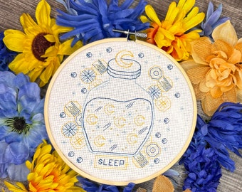 Può includere: Un cerchio da ricamo bianco con un disegno di una bottiglia di vetro con un tappo di sughero. La bottiglia è piena di stelle e le parole "SLEEP" sono scritte sotto la bottiglia. Il cerchio è circondato da fiori gialli, blu e bianchi.