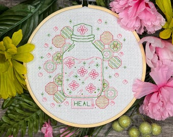 Può includere: Un cerchio da ricamo a punto croce con un disegno di un barattolo di vetro pieno di fiori rosa e la parola "HEAL" sotto. Il barattolo è circondato da forme di favo verde e arancio.