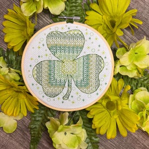 May include: A green shamrock cross-stitch design in a wooden embroidery hoop surrounded by yellow flowers.