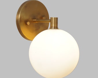 May include: A brass wall sconce with a white globe-shaped glass shade. The sconce has a round back plate and a simple arm.