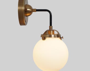 May include: A wall-mounted light fixture with a brass finish and a black curved arm. The fixture features a round, frosted glass globe. The design is reminiscent of vintage or industrial lighting.