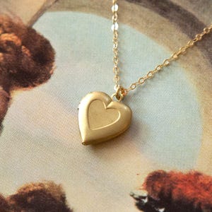 May include: A gold-colored heart-shaped locket necklace. The locket has a smaller heart design in the center and hangs from a delicate gold chain. The background is a vintage-style print with muted colors.