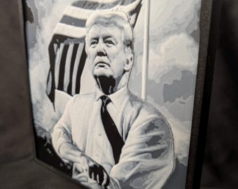 May include: A square, framed wall art piece featuring a grayscale, layered relief portrait of Donald Trump wearing a dress shirt and tie. Behind the figure, a stylized American flag is depicted with wavy, dimensional lines in shades of gray and white.