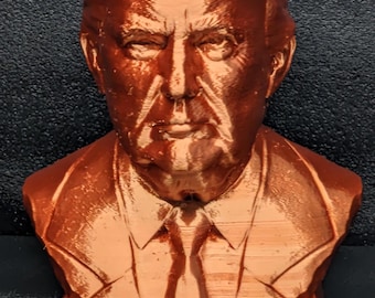 May include: A 3D printed bust of a person with a brown, textured finish. The bust is of a person with short, combed-over hair. The bust is on a base with the word "TRUMP" printed on it.