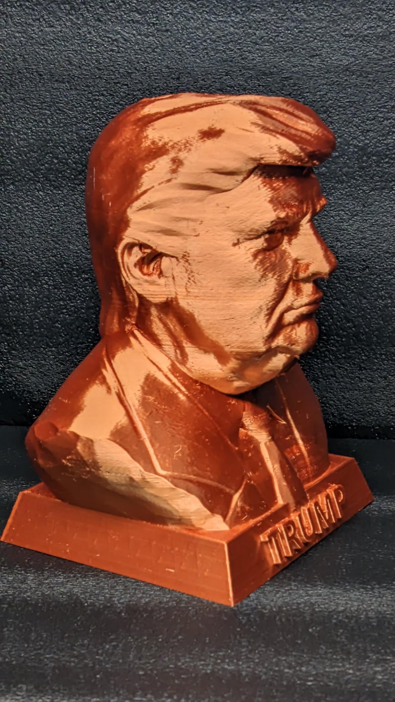 May include: A 3D printed bust of a person with the word "TRUMP" printed on the base. The bust is made of a brown plastic material.