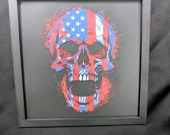 May include: A square black frame containing a three-dimensional skull art piece. The skull features a red, white, and blue American flag pattern with a distressed, splattered effect. The design is set against a solid black background.