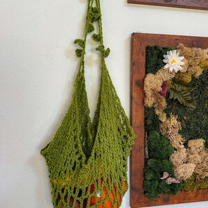Puede incluir: Una bolsa de mercado de ganchillo verde hecha a mano, colgada en una pared blanca. La bolsa está llena de naranjas y otras frutas. El asa de la bolsa está decorada con hojas de ganchillo. La bolsa está suspendida de un gancho blanco.