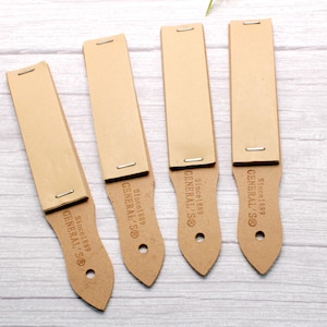 Puede incluir: Cuatro correas de cuero marrón con el texto "GENERAL S® Since 1889" impreso en el mango. Las correas tienen una superficie rectangular de cuero unida a un mango con un agujero en la parte inferior.