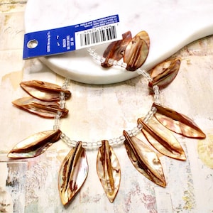 May include: A necklace featuring iridescent, leaf-shaped beads in shades of brown and gold, strung on a clear, beaded strand. The necklace is displayed on a white, curved surface. A blue tag with text is visible.
