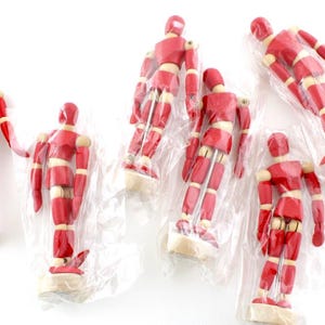 Op de afbeelding: Een verzameling rode houten kunstenaarsmannequins met beweegbare gewrichten, sommige individueel verpakt in doorzichtig plastic. De figuren staan op kleine, lichtgekleurde voetstukken. De achtergrond is wit.