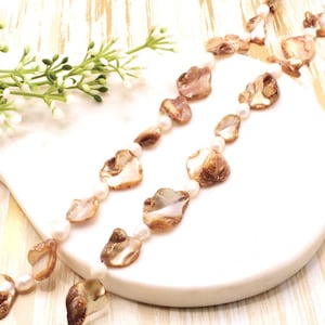 Op de afbeelding: Twee strengen van een ketting met onregelmatige, iriserende schelpstukken en kleine witte parels. De schelpen hebben bruine en crèmekleurige tinten. De ketting wordt gepresenteerd op een wit, gebogen oppervlak, met een takje witte bloemen.