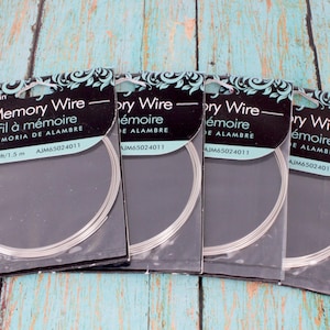 May include: Four packages of memory wire for jewelry making. Each package has a black and teal label with the words "Memory Wire" in English, French, and Spanish. The wire is silver and comes in a circular shape.