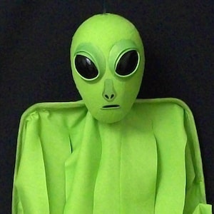 May include: A bright lime green alien figure featuring a smooth, rounded head with large, oval-shaped black eyes, a small nose, and a thin mouth line. The figure is draped in a matching lime green fabric that hangs in vertical folds.