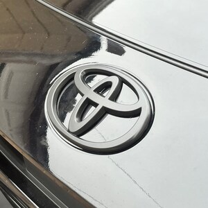 May include: Close-up of a black car hood with a silver Toyota emblem. The emblem is a stylized design of overlapping ovals, set against the glossy black surface. The hood reflects the surrounding environment.