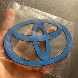 May include: A blue Toyota emblem in a clear plastic bag. The emblem has a black outline and a light blue interior. The Toyota logo is an oval shape with three overlapping ovals inside.