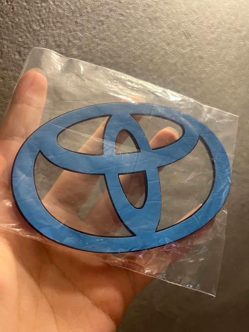 May include: A blue Toyota emblem in a clear plastic bag. The emblem has a black outline and a light blue interior. The Toyota logo is an oval shape with three overlapping ovals inside.