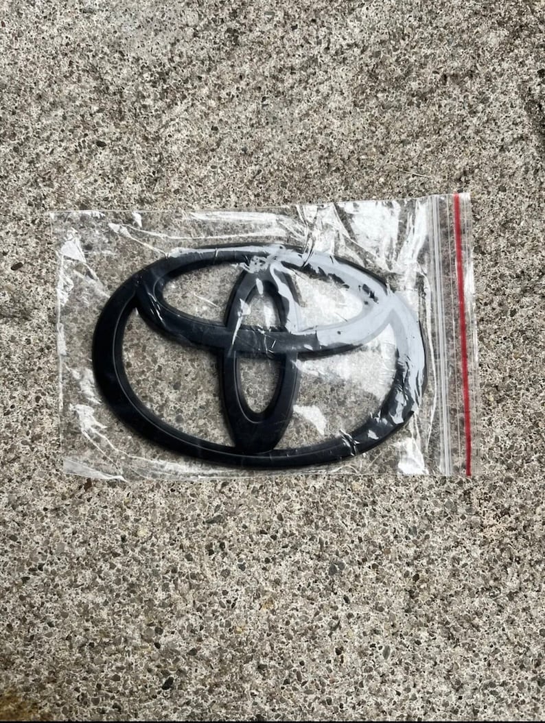 May include: A black Toyota emblem in a clear plastic bag. The emblem features the Toyota logo, a stylized oval design with overlapping inner shapes. The bag has a red zipper closure.