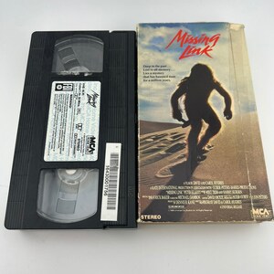Può includere: Una videocassetta VHS vintage e la sua custodia del film "Missing Link". La custodia presenta una figura silhouette su un paesaggio sabbioso. La videocassetta VHS è nera con etichette e testo bianchi. La custodia ha il logo MCA Home Video.