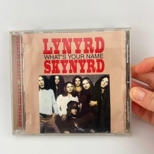 May include: A Lynyrd Skynyrd CD case featuring the album "What's Your Name." The album cover has a sepia tone with the band members' photo and the band's name in red. The CD is in a clear plastic case.