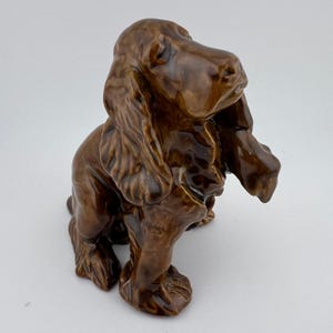 May include: A brown ceramic dog figurine with long, floppy ears and a detailed, textured coat. The dog is in a seated position, with its front paws extended. The figurine has a glossy finish and is set against a plain background.