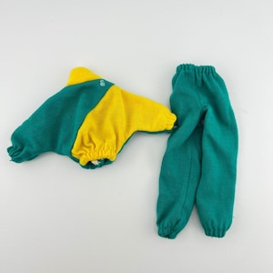 May include: A doll-sized outfit featuring a teal and yellow color block design. The top has long sleeves and a gathered waist. Matching teal pants with an elastic waistband complete the set. The outfit is made of a soft, knit fabric.