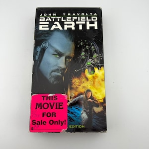 May include: A VHS tape for the movie "Battlefield Earth" starring John Travolta. The cover features a close-up of Travolta's face with green eyes, and a scene of a man running. A pink sticker reads "THIS MOVIE FOR Sale Only!"