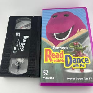 May include: A black VHS tape and its corresponding purple-edged plastic case. The case features a purple dinosaur character, a green character, and the text Barney's Read with Me Dance with Me, 52 Minutes, and Never Seen On TV. The tape has a label.