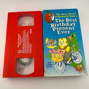 May include: A red VHS tape and a book titled "The Best Birthday Present Ever" from "The Busy World of Richard Scarry." The book cover is blue with illustrations of cartoon characters and a bicycle.