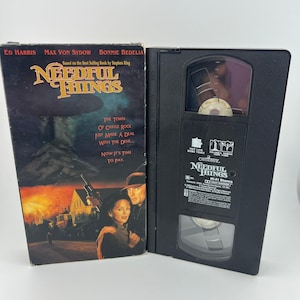 May include: A VHS tape of the movie "Needful Things" is displayed open. The cover art features Ed Harris, Max Von Sydow, and Bonnie Bedelia. The title is in a gold, stylized font. The tape is black with the title and other text.