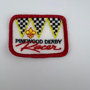 May include: Embroidered patch with a red border and a white center. The patch features the text "Pinewood Derby Racer" in red and black. Above the text are three yellow-bordered pennants with black and white checkered patterns. A gold fleur-de-lis is centered.