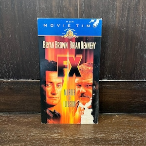 May include: A VHS tape for the movie "FX Murder by Illusion" featuring Bryan Brown and Brian Dennehy. The cover has a blue top with the MGM Movie Time logo, and a fiery background with the movie title and actors' names.