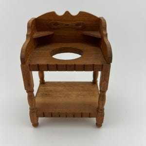 May include: A miniature wooden dollhouse sink with a round basin and a lower shelf. The sink has a decorative top with a carved design and is made of a light brown wood. The legs are turned and the overall design is vintage.