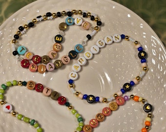 May include: Four beaded bracelets with different colored beads and gold letter charms spelling out the words "LOVE", "CARDIGAN", "AUGUST", and "BETTY".