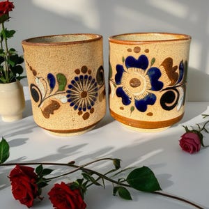 May include: Two cylindrical ceramic cups with a speckled beige finish, each featuring hand-painted floral motifs in deep blue, brown, and green. The cups have a smooth, rounded rim and a slightly tapered base, showcasing intricate petal and leaf patterns.